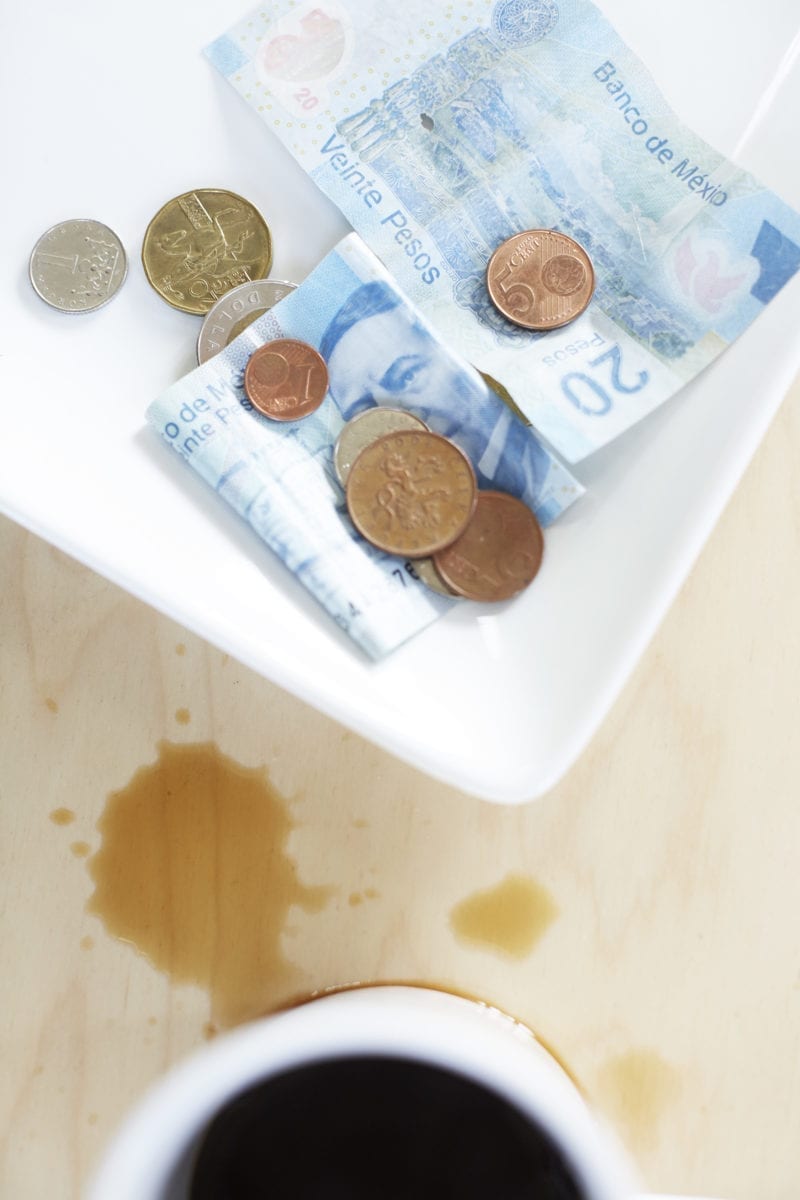 A cup of coffee with some spilled on the table and a bowl of coins