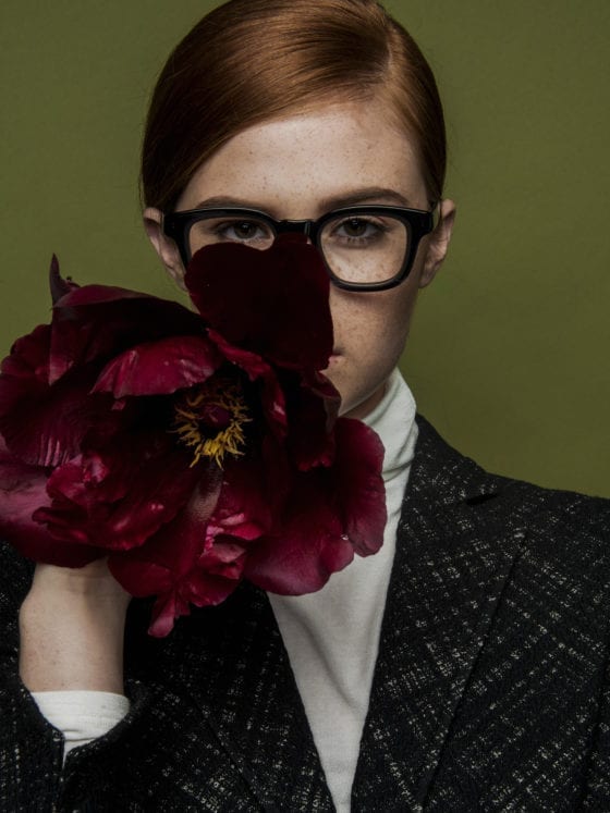 A red-headed woman in a suit jacket with black, plastic glasses and a white shirt, holding red flowers