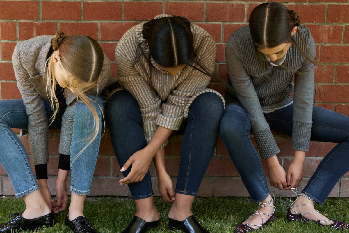 Three women looking down at their feet while tying their shoes along a brick wall