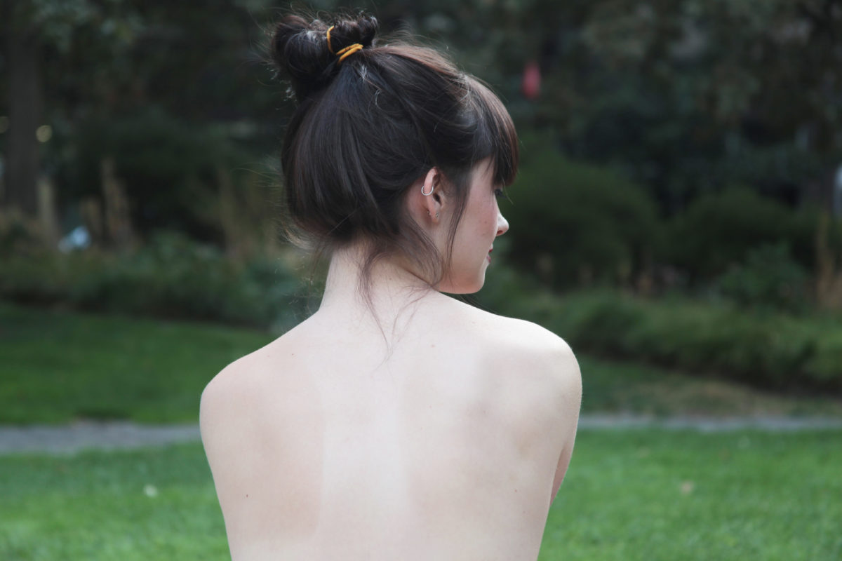 The back of a topless woman with her hair in a bun and facing away from the camera as she sits outside