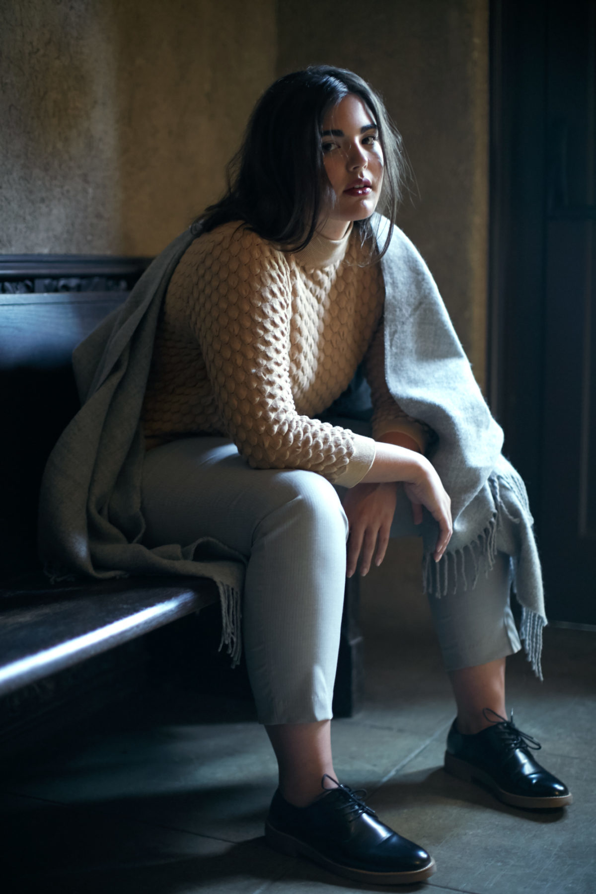 A woman sitting on a bench inside a dark room with a shawl wrapped around one shoulder, a sweater and denim pants