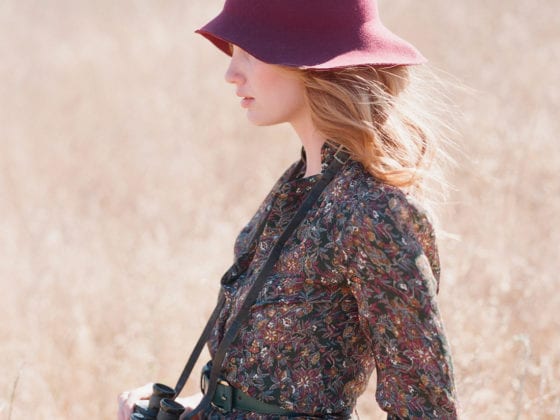 A woman with a hat, long-sleeved top and skirt standing in a field with binoculars