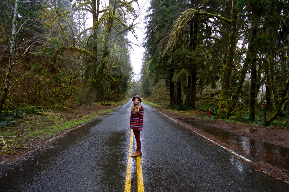 A woman standing in the middle of a road surrounded by trees on a rainy day