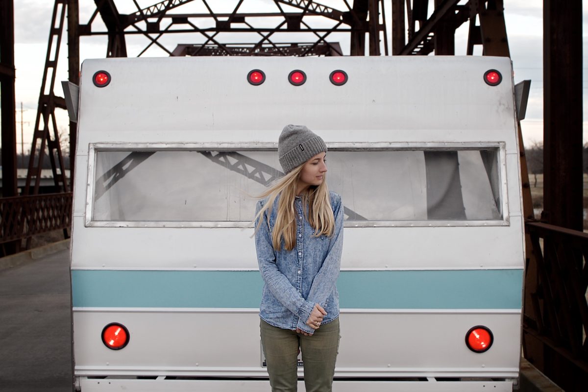 A woman with long hair wearing a beanie hat, jean jacket and pants standing in front of a trailer home