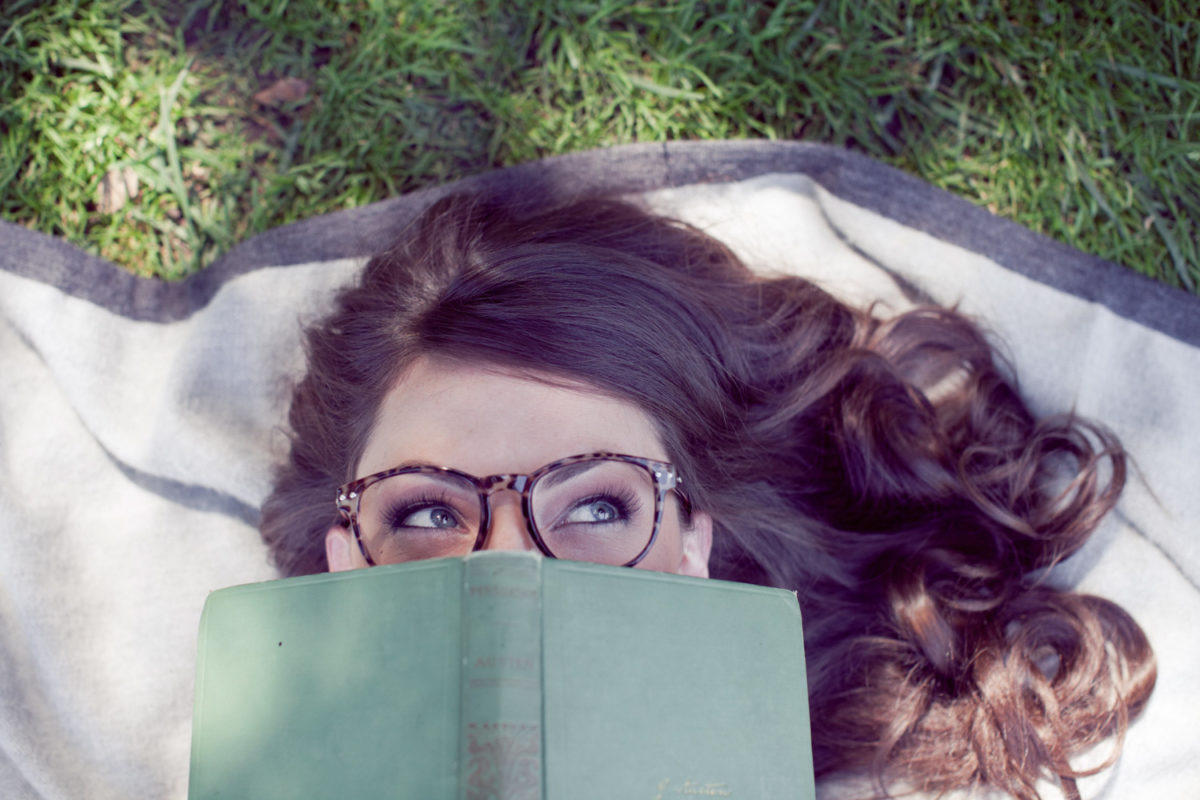 An unclose picture of a girls with glasses holding a book to her face as she lies on a blanket outside