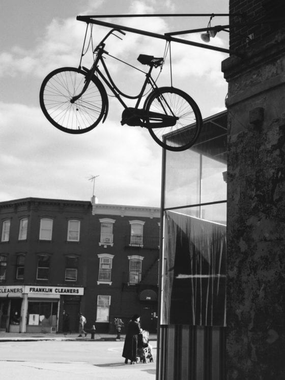 A black and white photo of a city street with a bicycle hanging from a wire