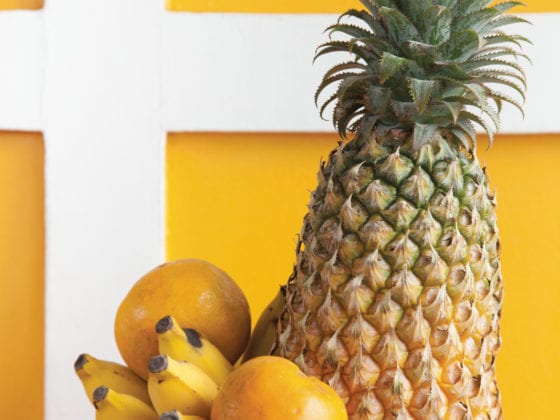 A photo of a fruit bowl with pineapple, banana and melon in front of a yellow wall