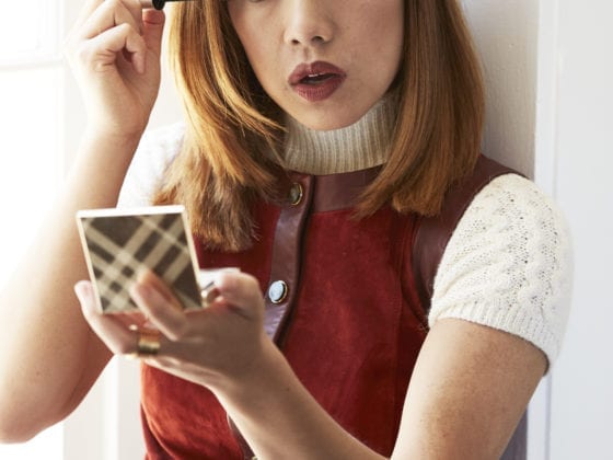 A woman putting on makeup as she looks into a compact mirror and sits near a window sill