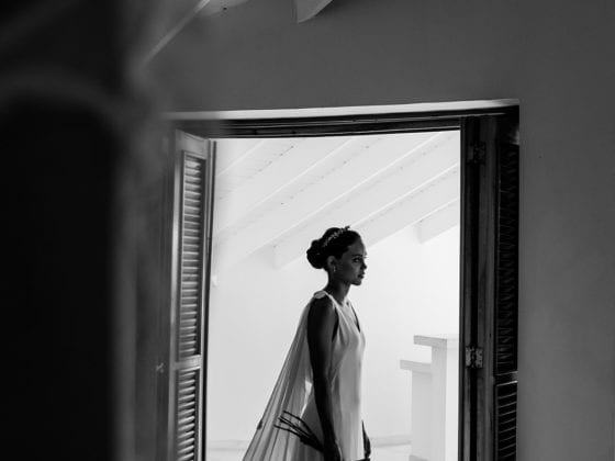 A black and white photo of a woman in her wedding gown and veil standing in front of a door that leads to outside