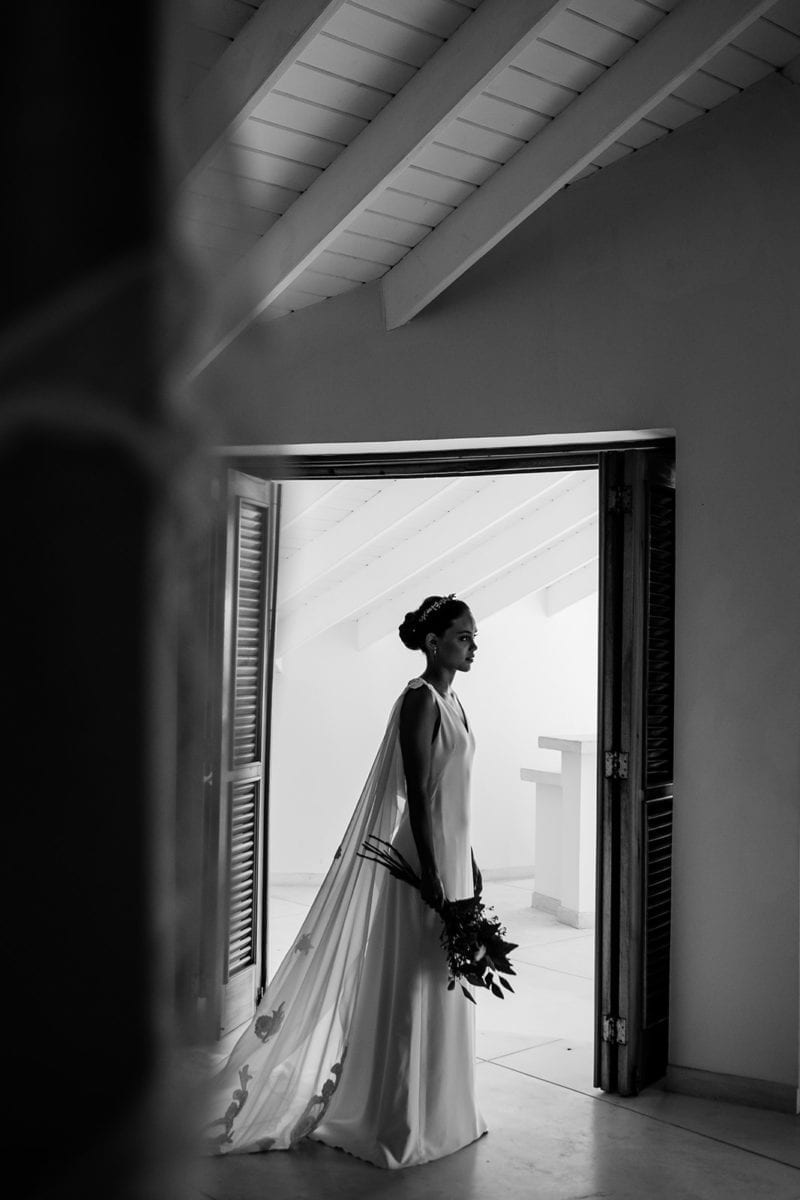 A black and white photo of a woman in her wedding gown and veil standing in front of a door that leads to outside