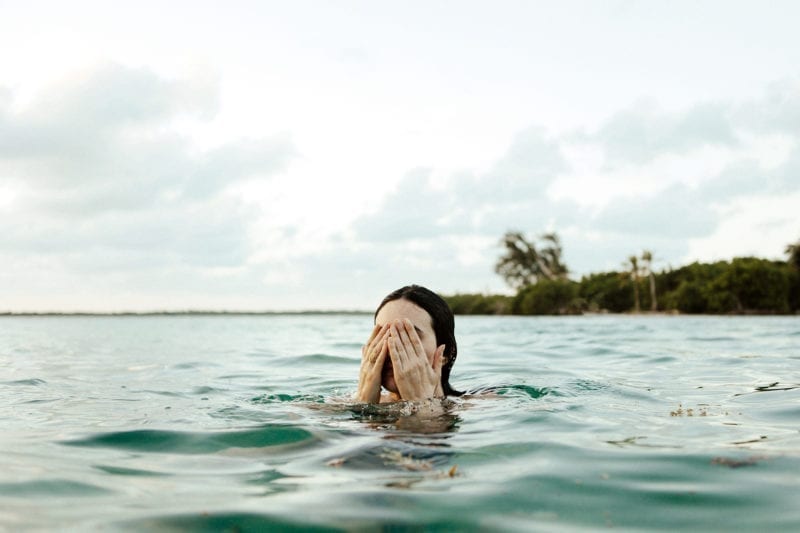 A woman in the ocean, fully submerged except for her head, with her hands wiping her eyes