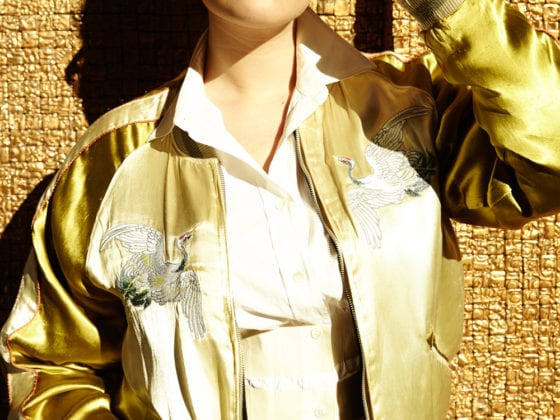 A woman in a gold jacket leaning against a gold wall with her sunglasses on her face