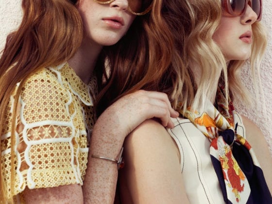 Two girls wearing sunglasses and one leans over the other's shoulder
