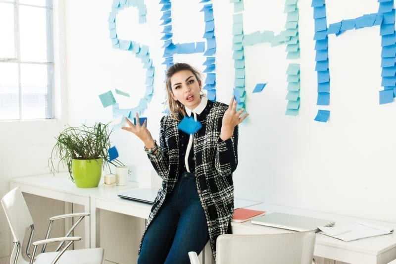 A woman seated on the edge of her desk with stickie notes that say, "Shhh!" on the wall behind her.