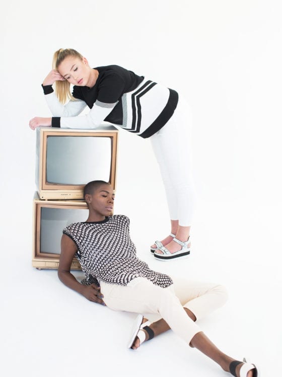 Two women leaning against two old school televisions that are stacked atop each other