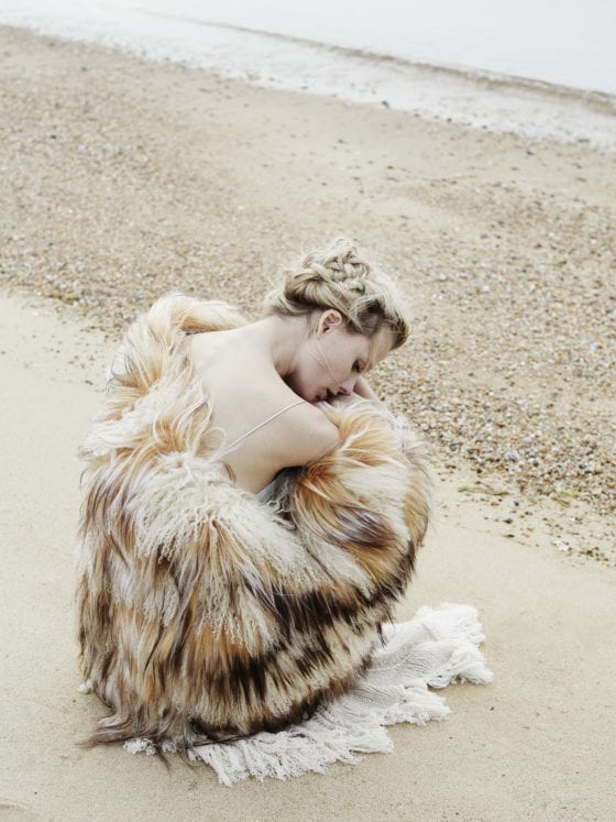 A woman with fur jacket falling off her shoulder as she crouches in the sand near a beach