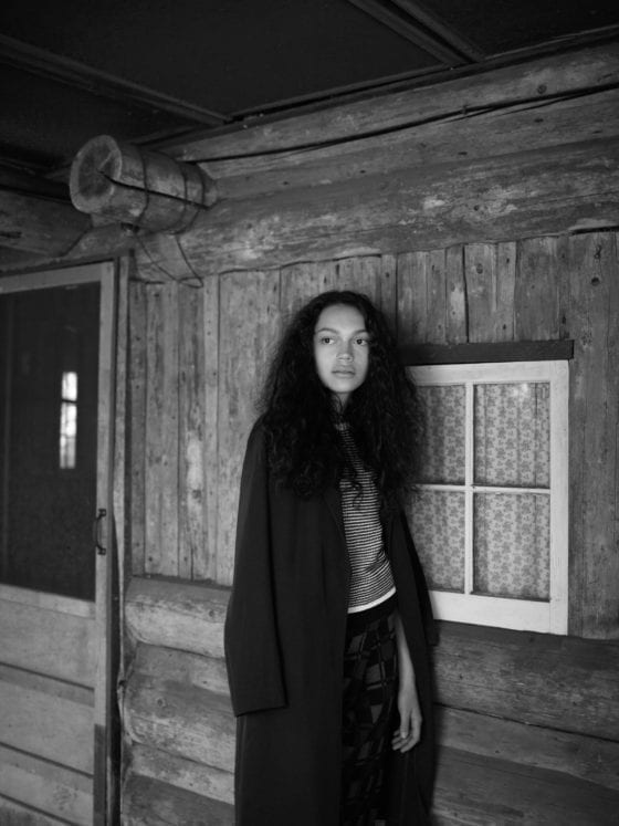 A black-and-white photo of a black woman with long hair and a black jacket standing outside of a cabin