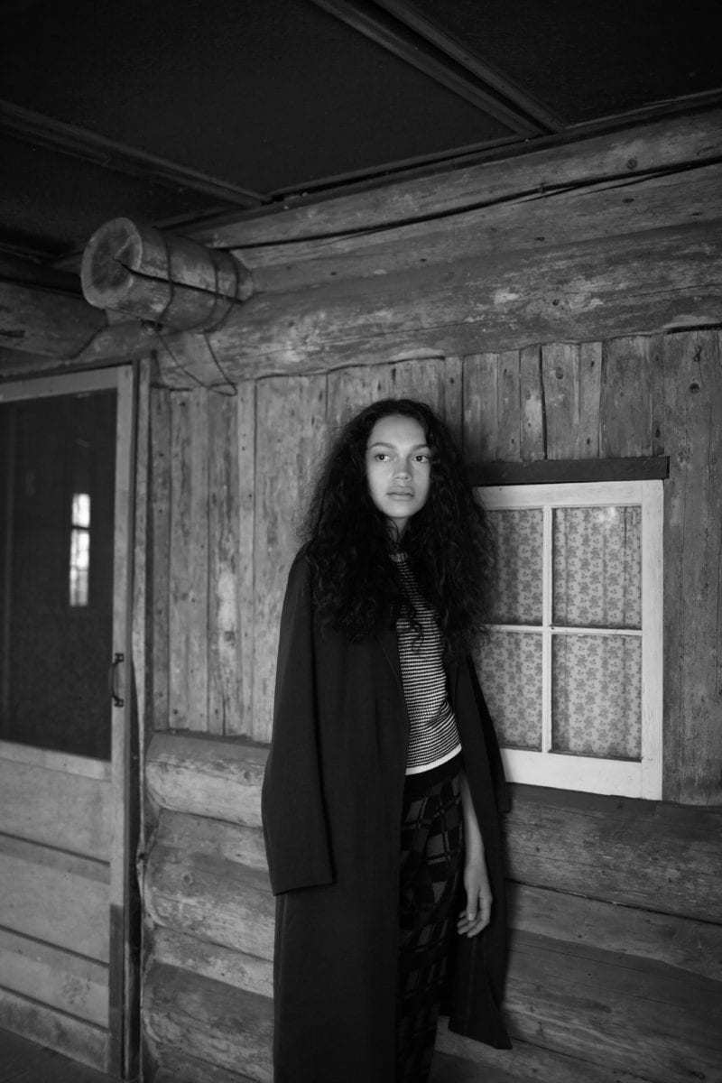 A black-and-white photo of a black woman with long hair and a black jacket standing outside of a cabin