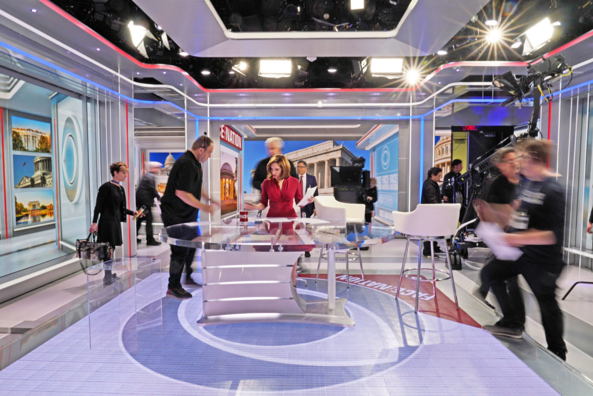 A news anchor stands at her desk while a film crew moves around her (out of focus)