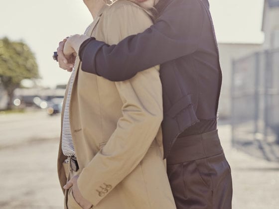 A woman leaning on the back of a man as she embraces him in a hug. Both are straight faced