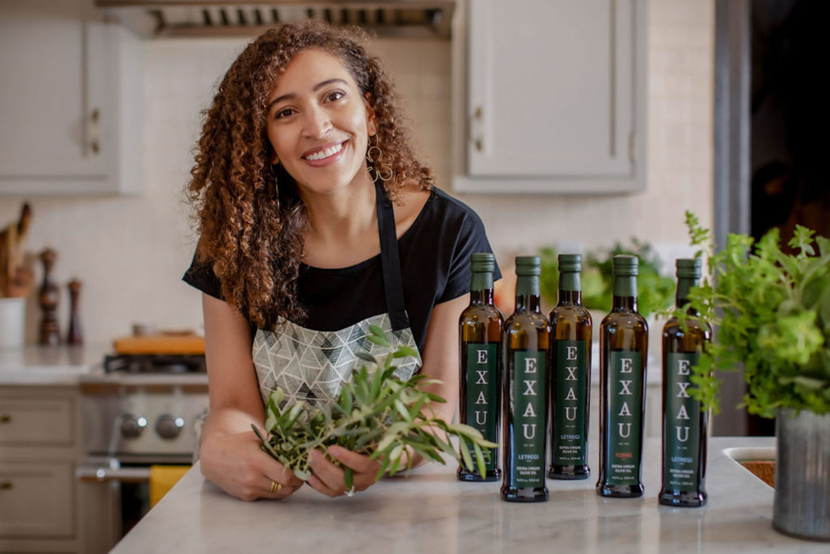 A woman olive an olive branch as she leans over a kitchen counter with olive oil bottles next to her