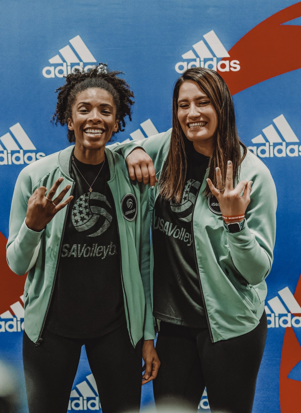 Two smiling women standing in front of an Adidas poster each gesturing three figures