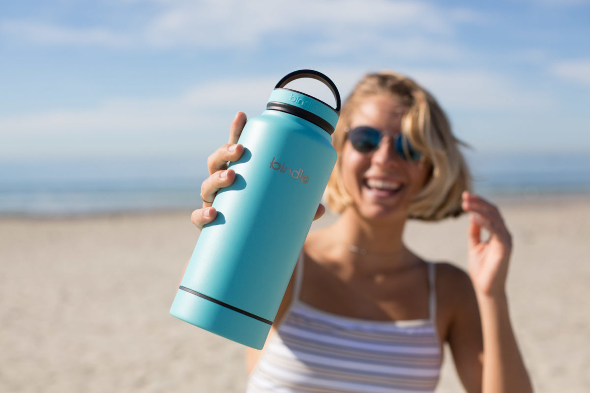 A smiling woman on a beach holding up a bottle toward the camera