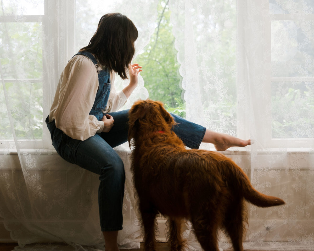 A woman wearing denim overalls and a blouse as she looks out of curtains and her dog looks on next to her