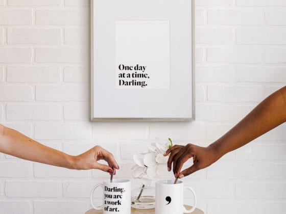 Two hands stirring coffee cups with a piece of art on the wall that says, "One day at a time, Darling."