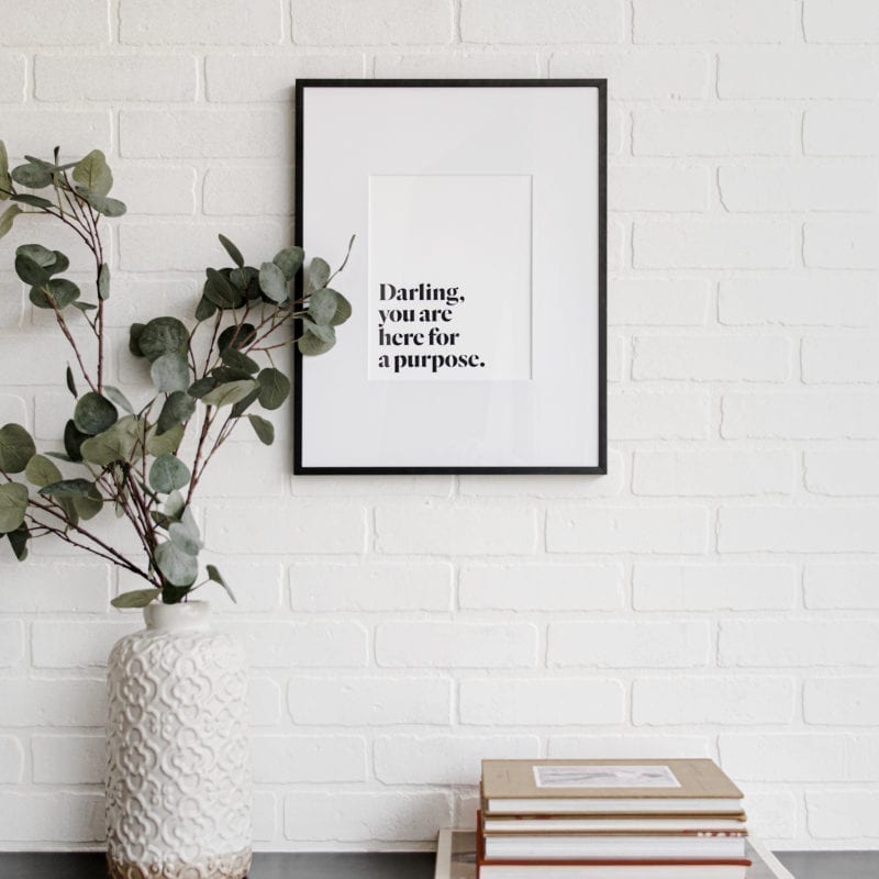 A picture of a piece of wall art that says, "Darling, you are here for a purpose" atop a book shelf with a stack of books and a greenery