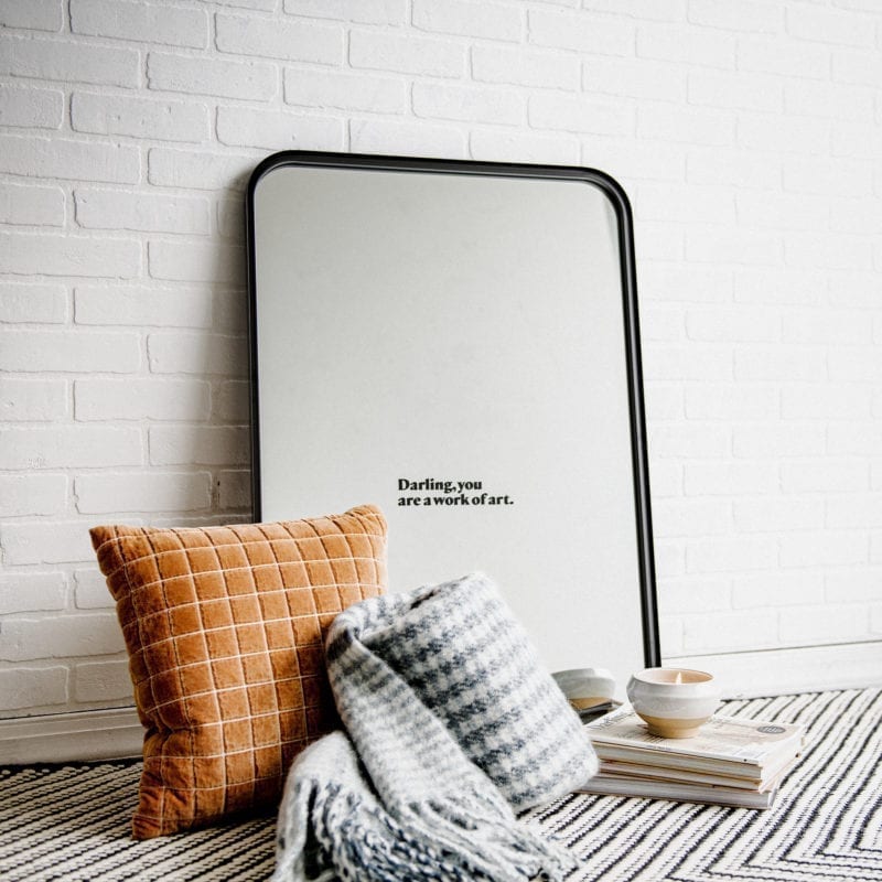 A mirror leaning against a wall with a pillow, throw blanket and a pile of books on the floor
