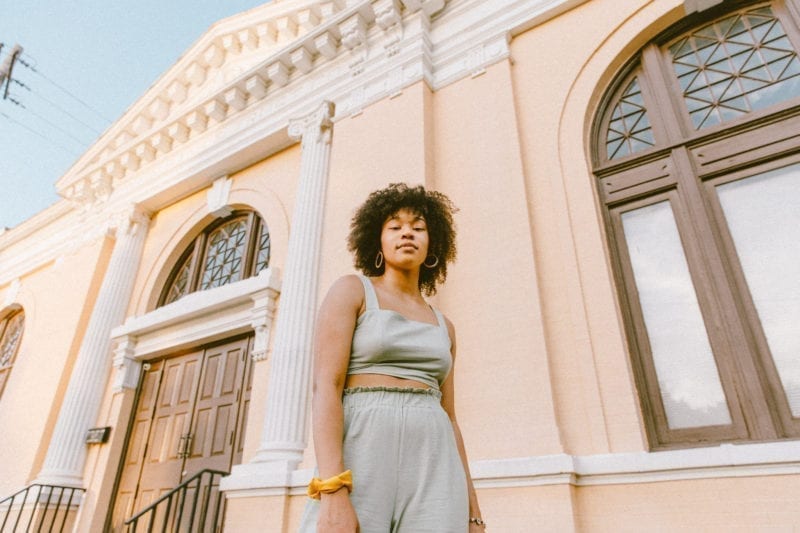 A picture of a black woman with curly hair standing outside of a pink building