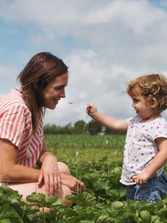 A mom and a daughter standing in a field as the baby girl holds a flower up to her moms face