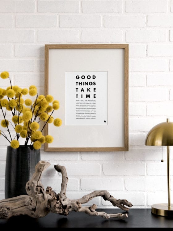 A piece of art that says "Good Things Take Time" near a table with a lamp and flowers
