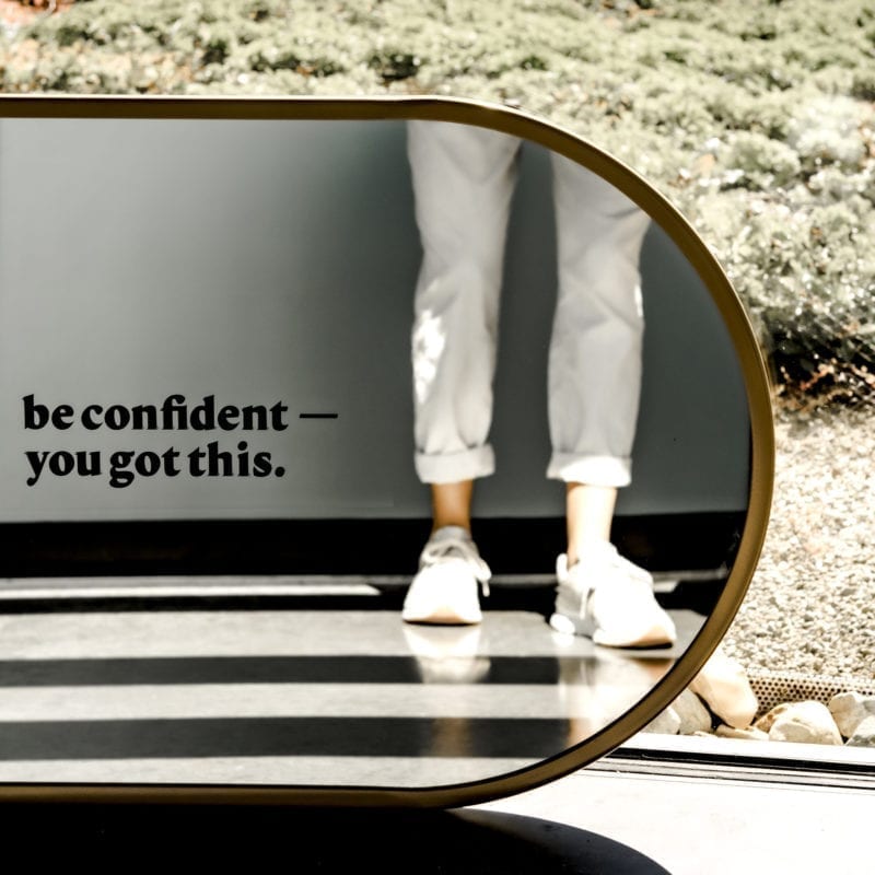 A woman's reflection in a mirror that has the decal "be confident--you got this."