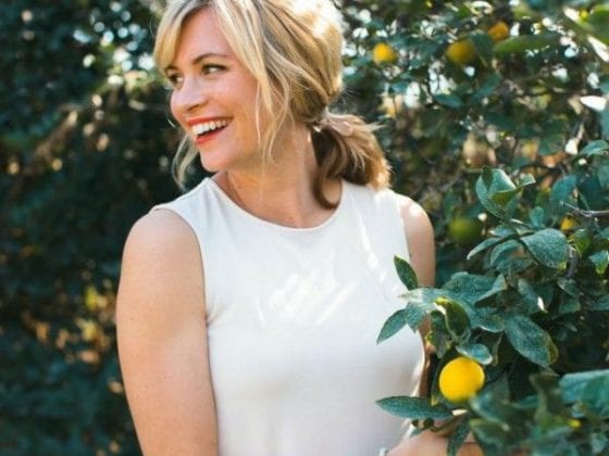 A smiling woman looking over her shoulders as she stands next to a lemon tree