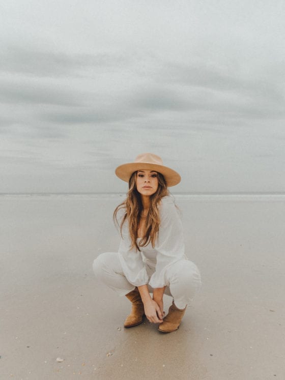 A woman with a fedora and white outfit crouched low outside at the beach