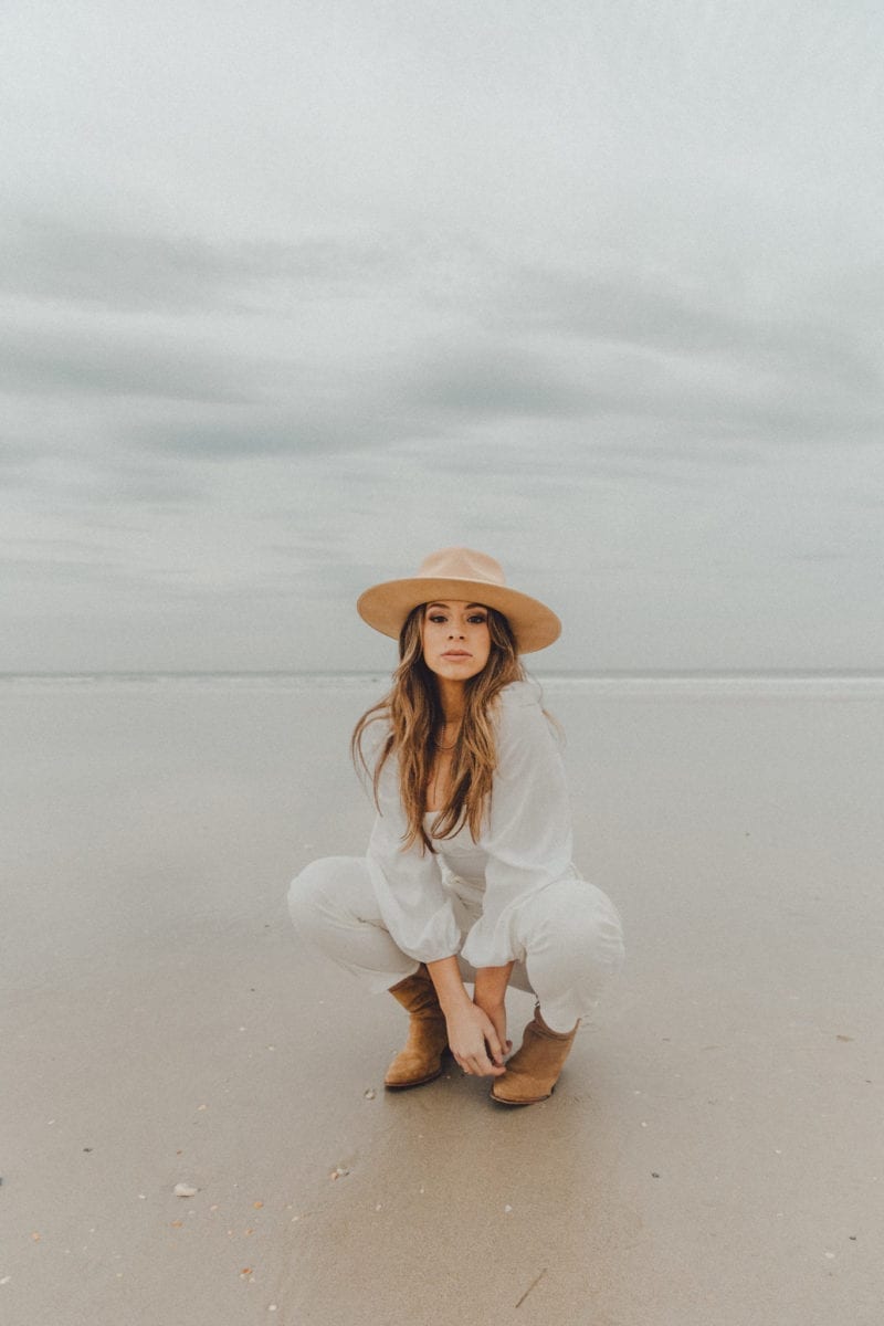 A woman with a fedora and white outfit crouched low outside at the beach