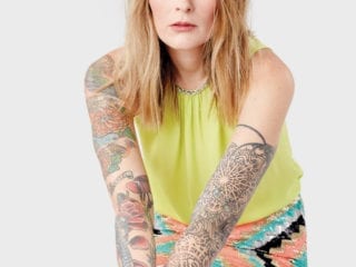 A woman with blondish red hair and arm tattoos looking straight into the camera