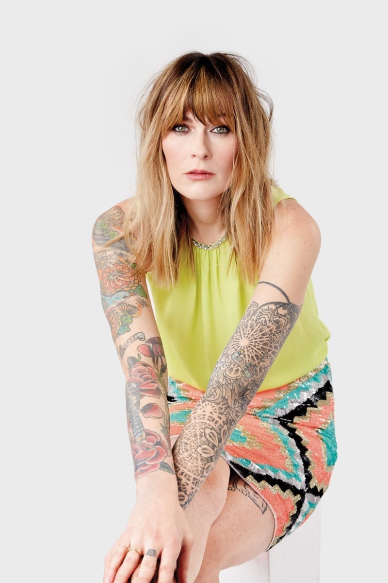 A woman with blondish red hair and arm tattoos looking straight into the camera