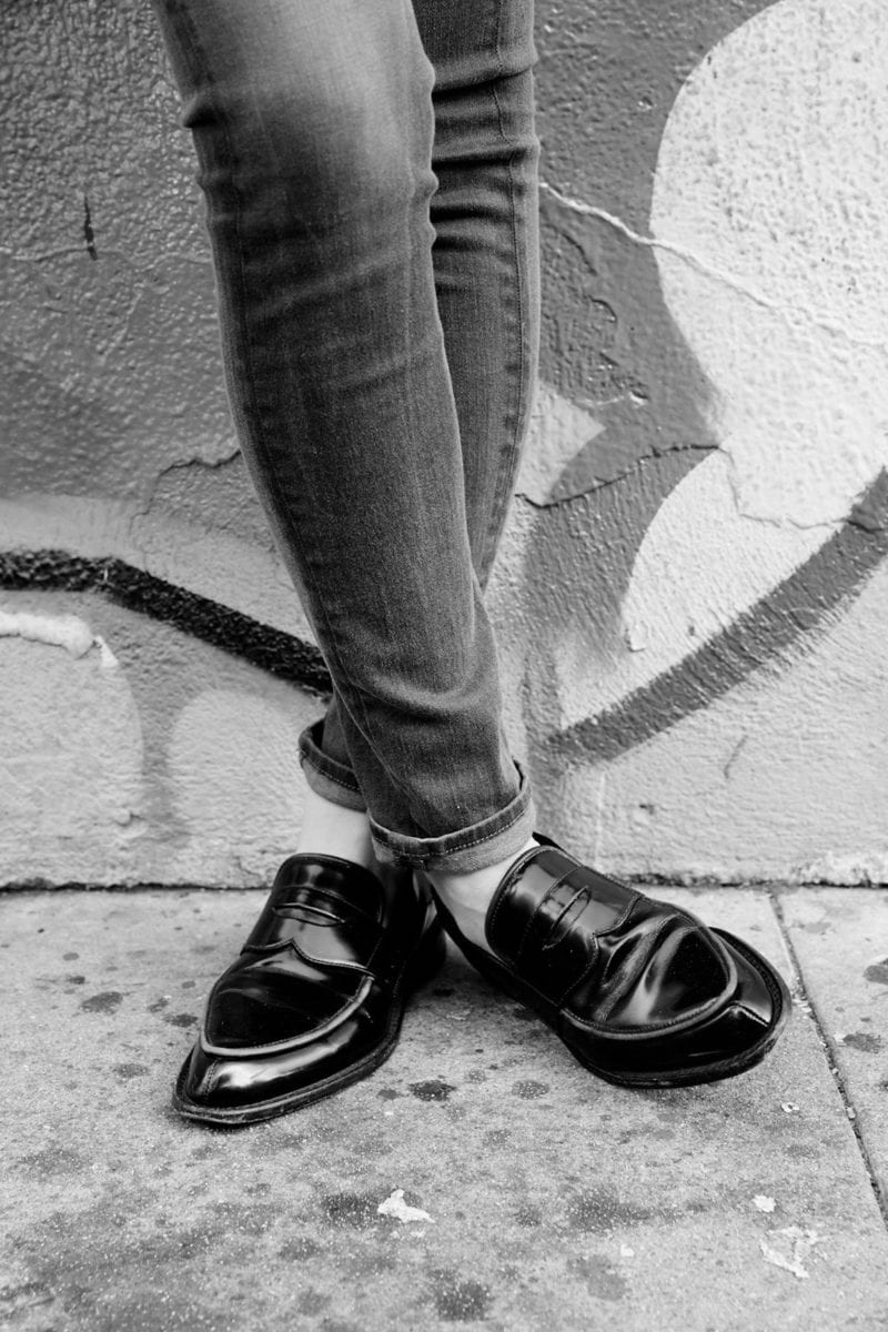 A black and white image of a woman's feet crossed as she stands outside on a sidewalk