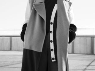 A black and white photo go a woman standing in a trench coat atop of a building